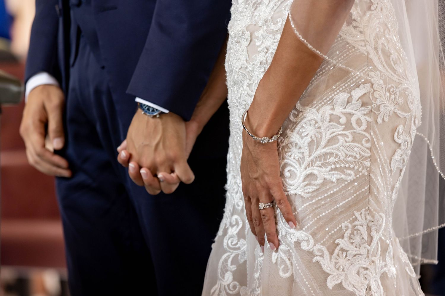 Close up view of a bride and groom holding hands, with lace wedding dress and navy suit details visible.