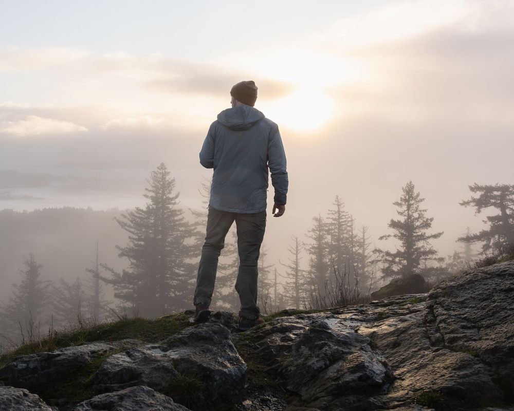 A silhouetted figure stands on a rocky mountaintop overlooking a misty forest landscape at sunset.
