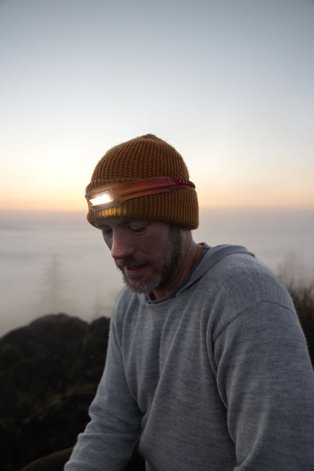 Person wearing a brown beanie and grey sweatshirt at sunset on a mountain peak.