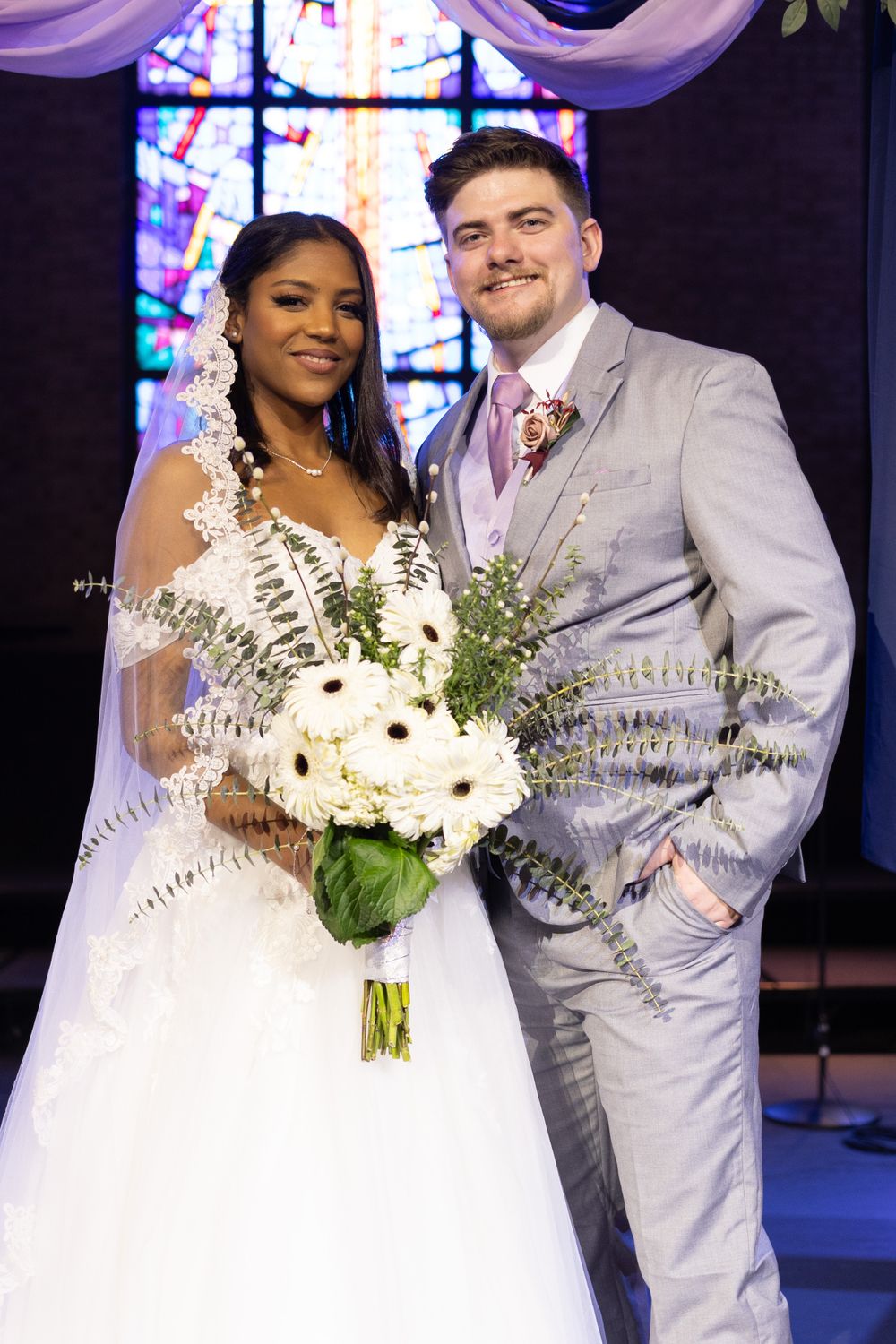 A wedding photo shows newlyweds posing together in front of a stained glass window, with the bride holding white flowers.