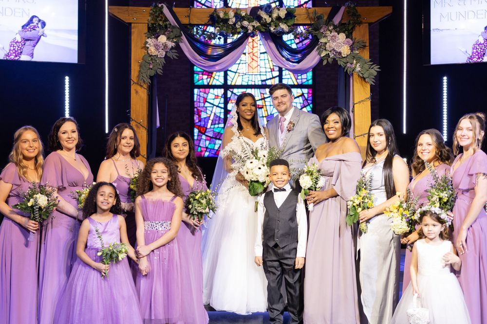 Wedding party photo with bridesmaids in purple dresses and lavender gowns standing together under decorative arch.
