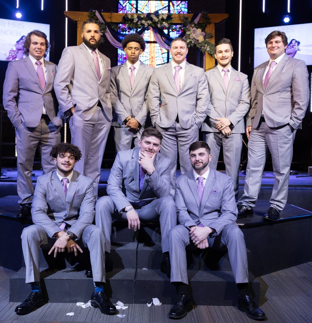 Groomsmen in matching gray suits pose together on stage with dramatic lighting and stained glass backdrop.