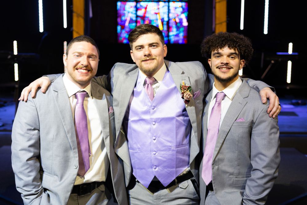 Three groomsmen in gray suits and pink ties pose together in front of church altar.