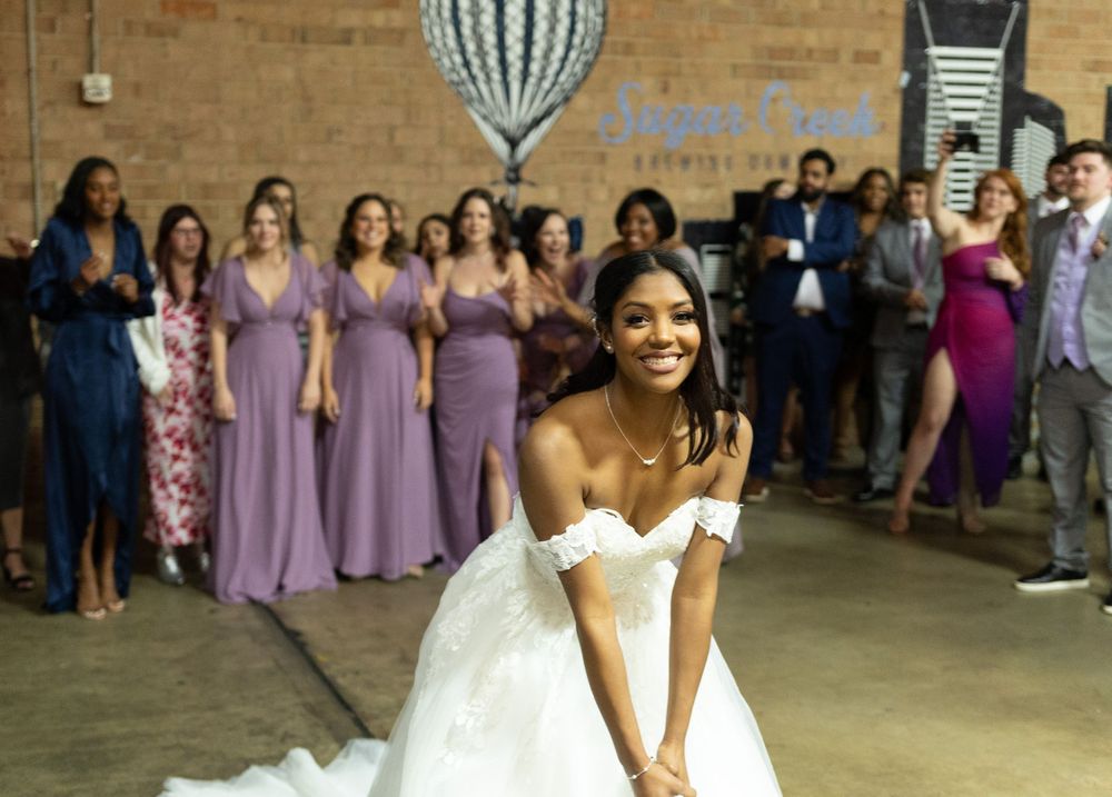 Bride in white gown smiling joyfully on dance floor with wedding party in purple dresses in background.