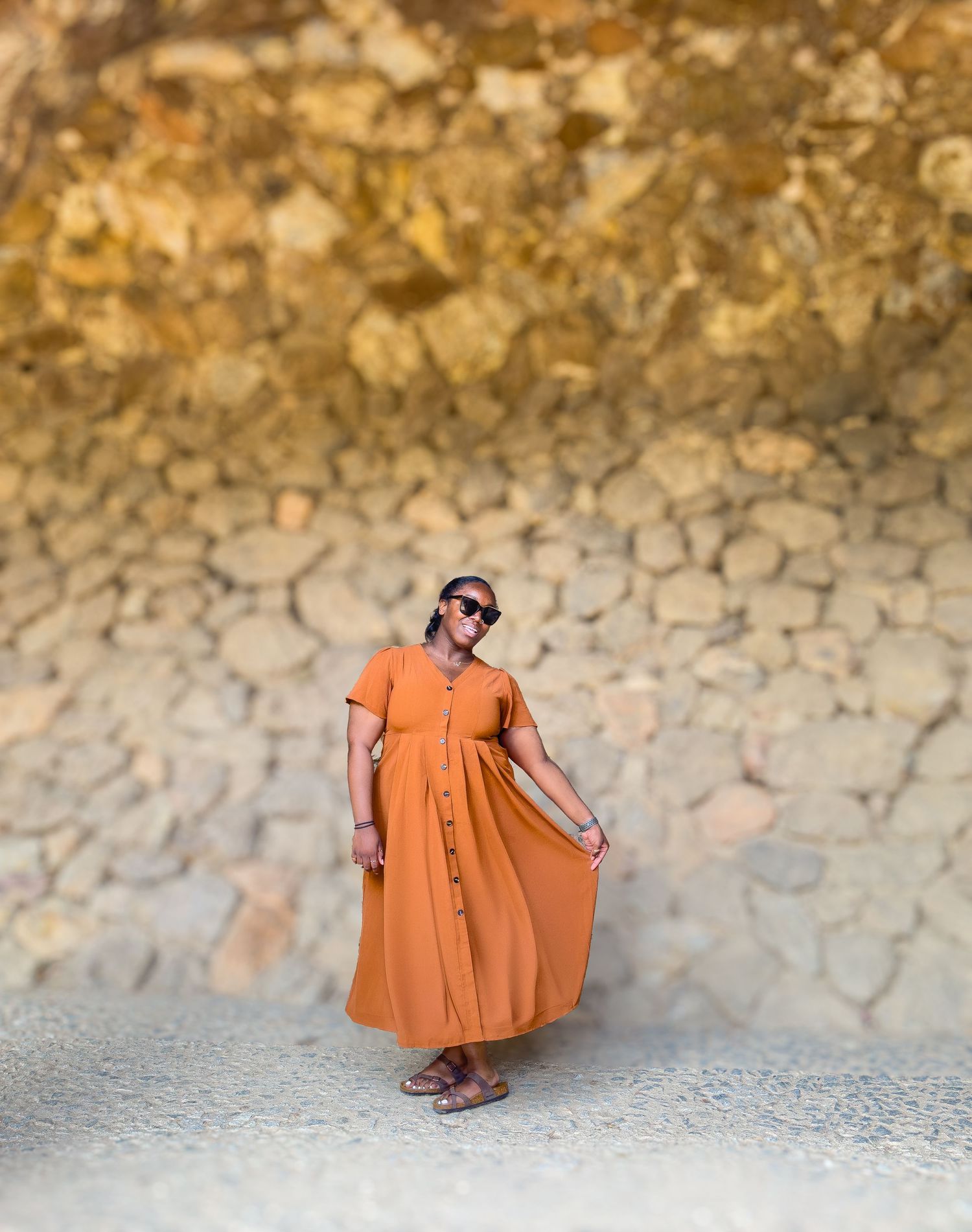 Someone wearing an orange button-down maxi dress and sunglasses poses against a textured stone wall outdoors.