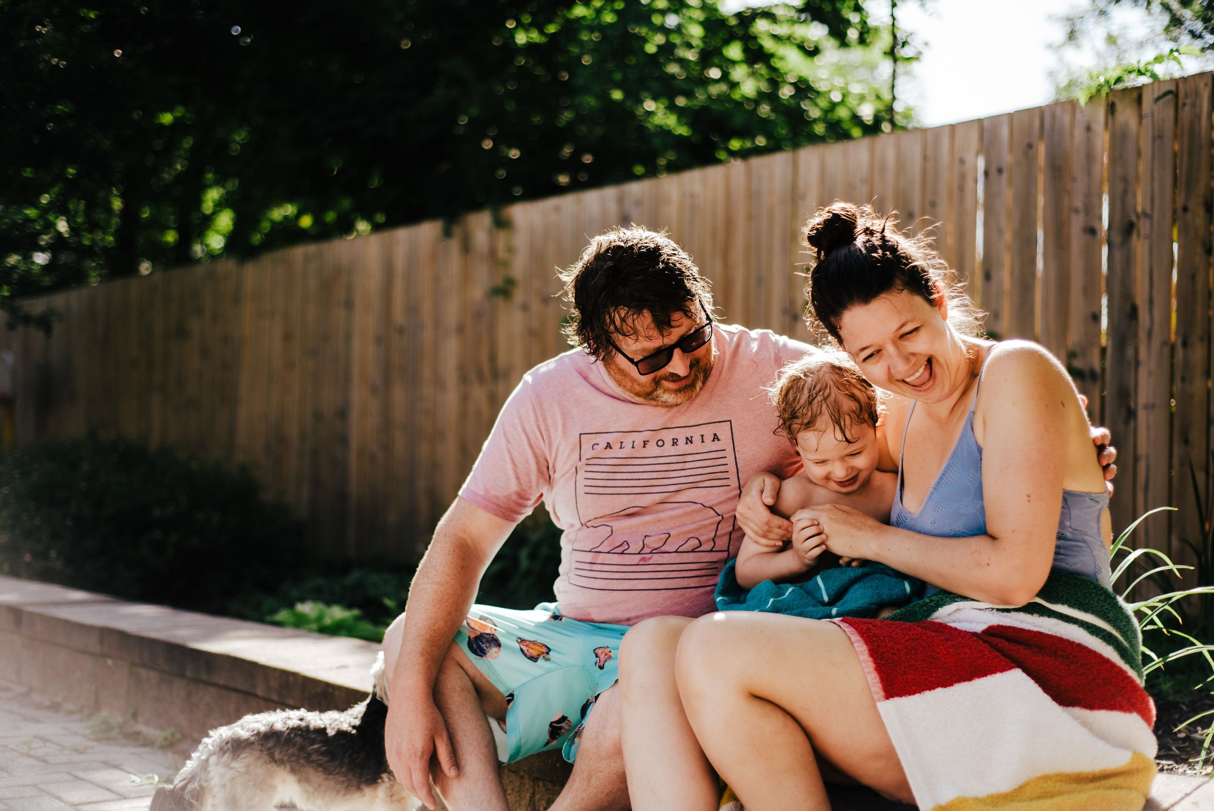 Three people sit together on a sunny summer day against a wooden fence in casual attire.