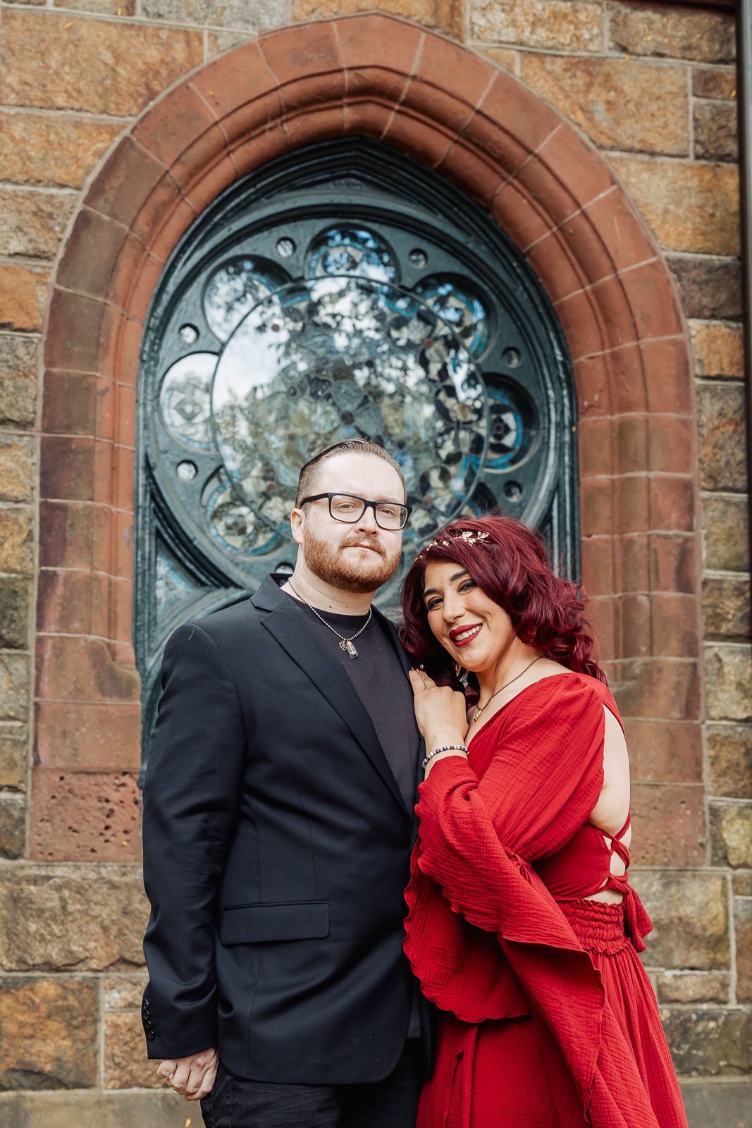 A couple poses in front of an ornate church doorway, with the woman wearing a striking red dress.