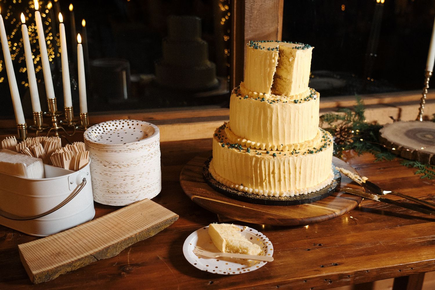 A three-tiered yellow buttercream wedding cake sits on a wooden table beside lit candles and table decorations.