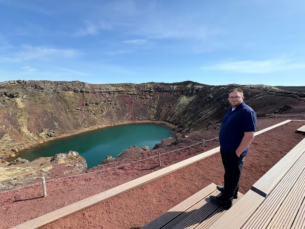 Person stands on a wooden platform overlooking Kerid Crater lake with turquoise water and red volcanic rocks in Iceland.