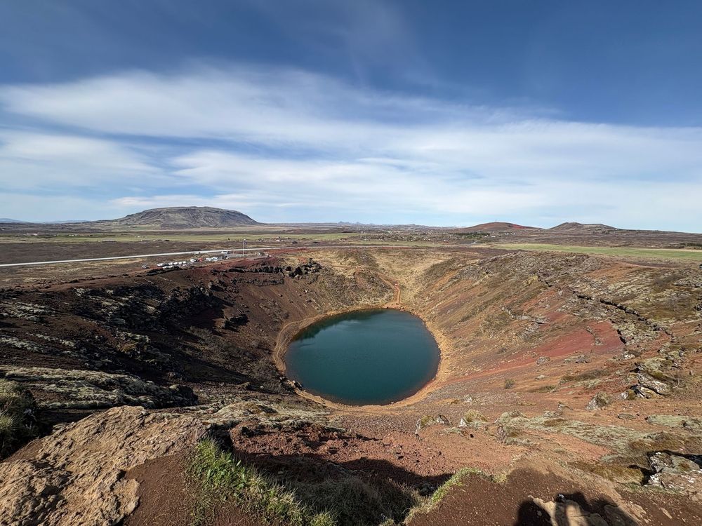 Aerial view of a circular volcanic crater lake with turquoise water surrounded by red volcanic rock formations in a desert landscape.