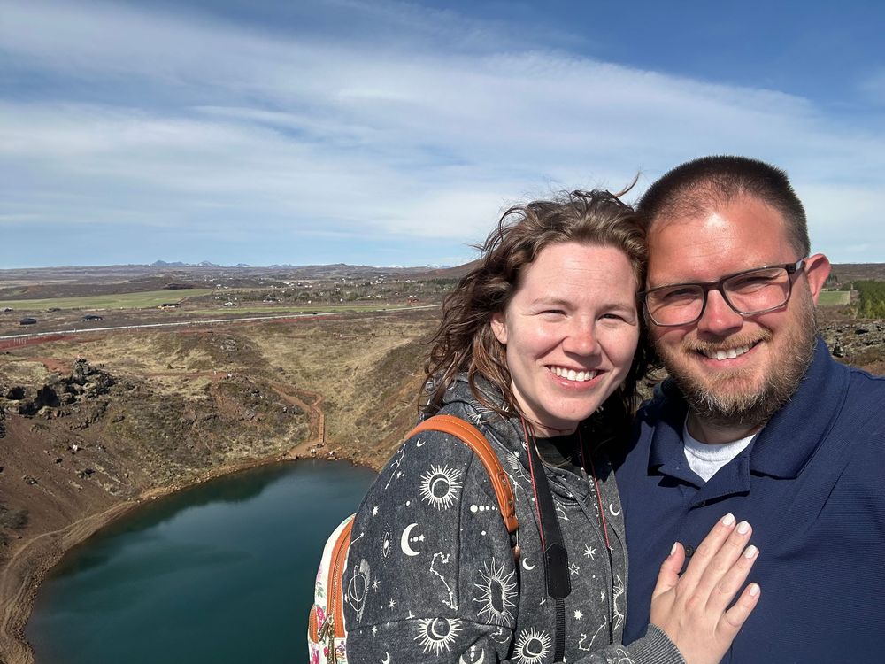 A couple takes a selfie overlooking a scenic blue crater lake with mountains and desert landscape in the background.
