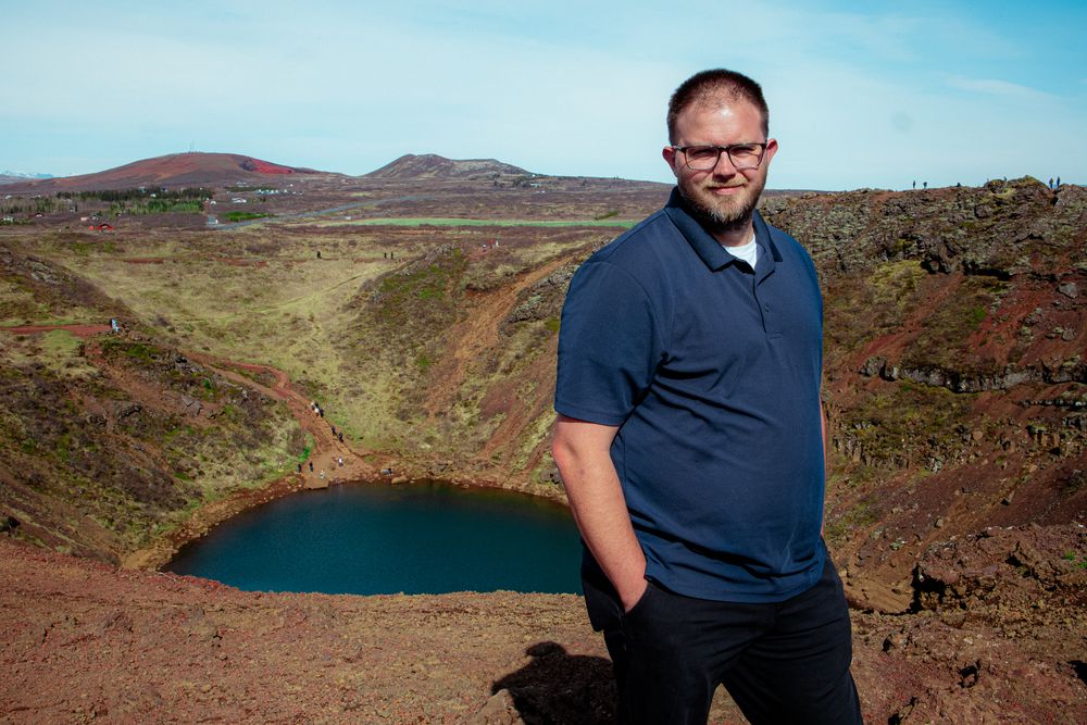 A visitor stands at the edge of a volcanic crater lake with turquoise waters and reddish volcanic terrain in Iceland.