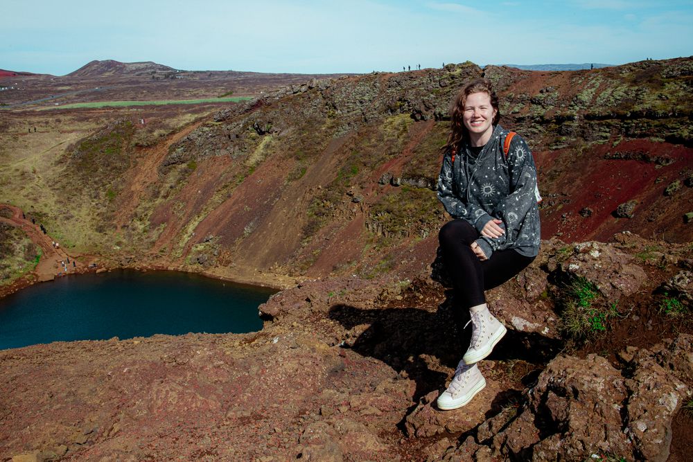 A person sits on the edge of a volcanic crater lake with red rocks and hills in the background in Iceland.