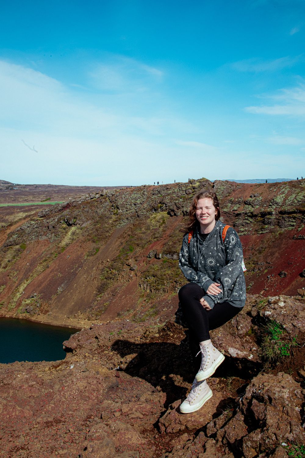 Tourist sits on the red rocky rim of a volcanic crater lake with blue sky above, creating a stunning landscape view.
