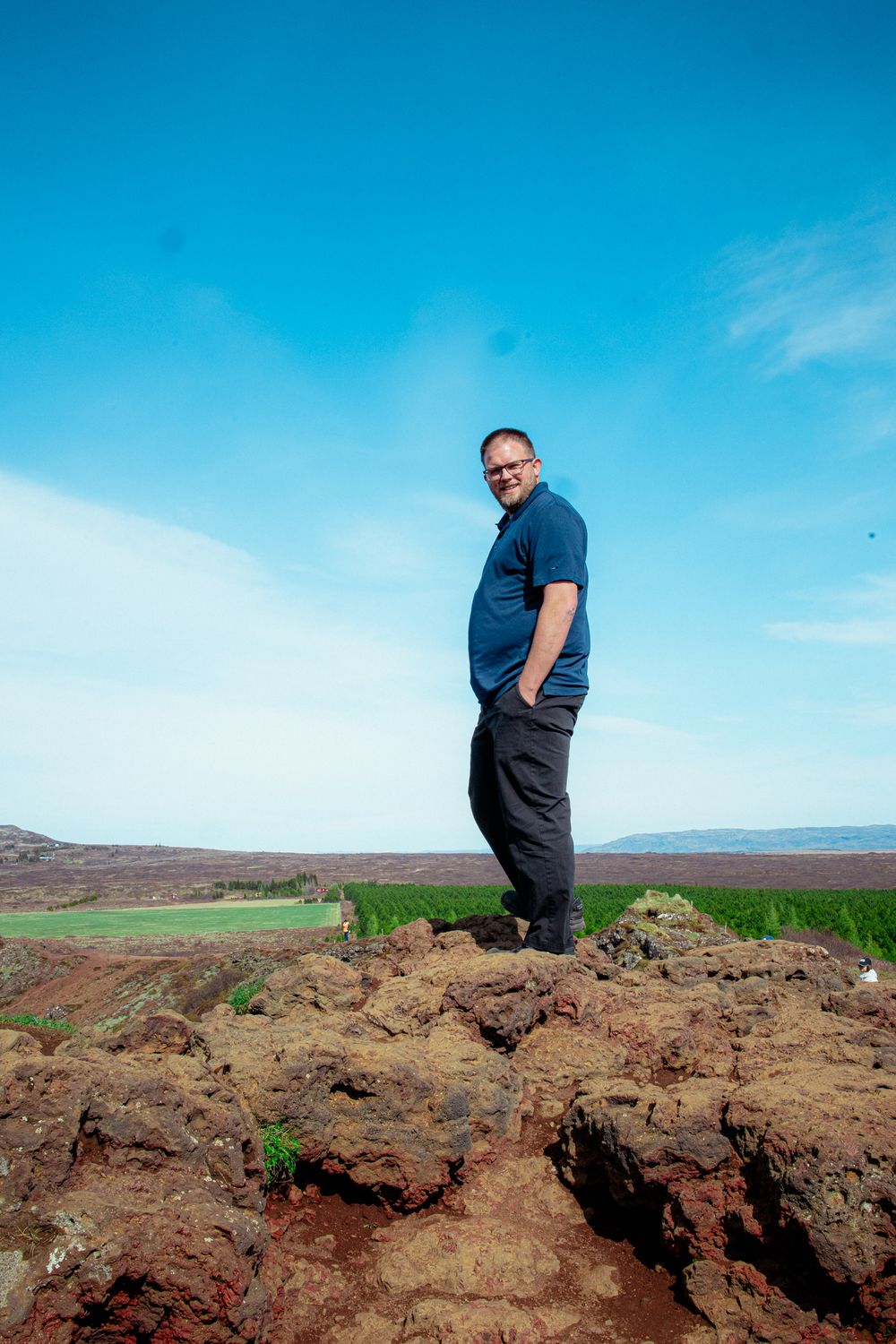 A figure stands on rocky terrain overlooking green rolling hills against a bright blue sky with wispy clouds.