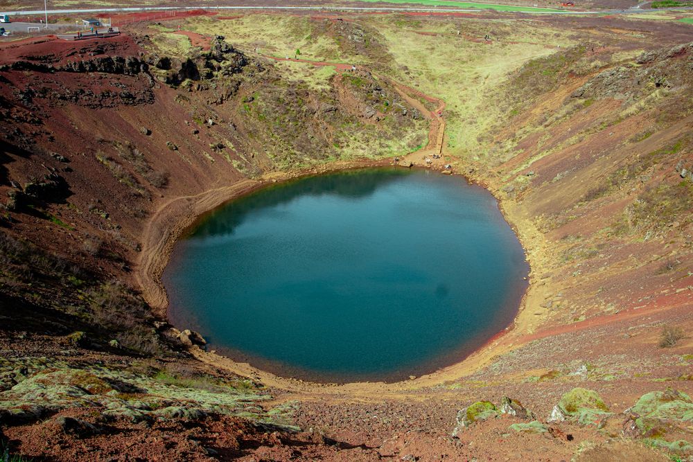 A circular turquoise crater lake surrounded by red-brown volcanic rock and green moss in the Icelandic highlands.