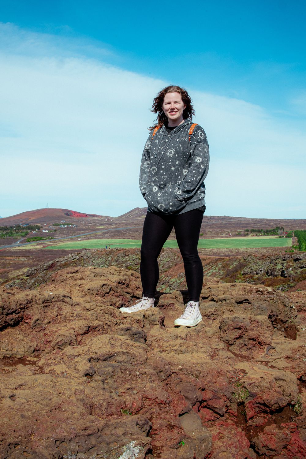 Person in a patterned sweater and dark pants stands on rocky terrain with green hills and blue sky in the background.