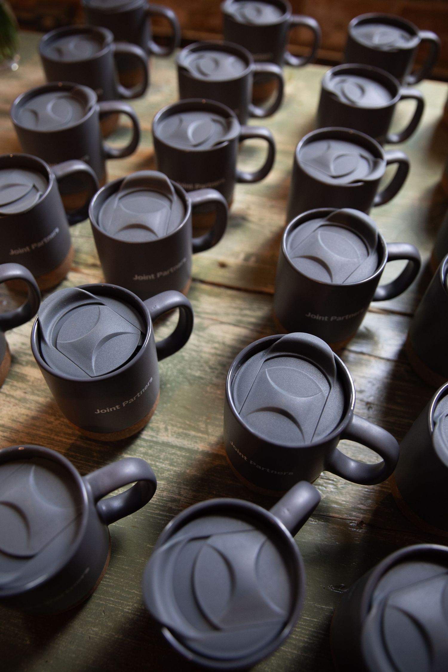 Multiple dark matte coffee mugs with lids arranged on a wooden surface in uniform rows.