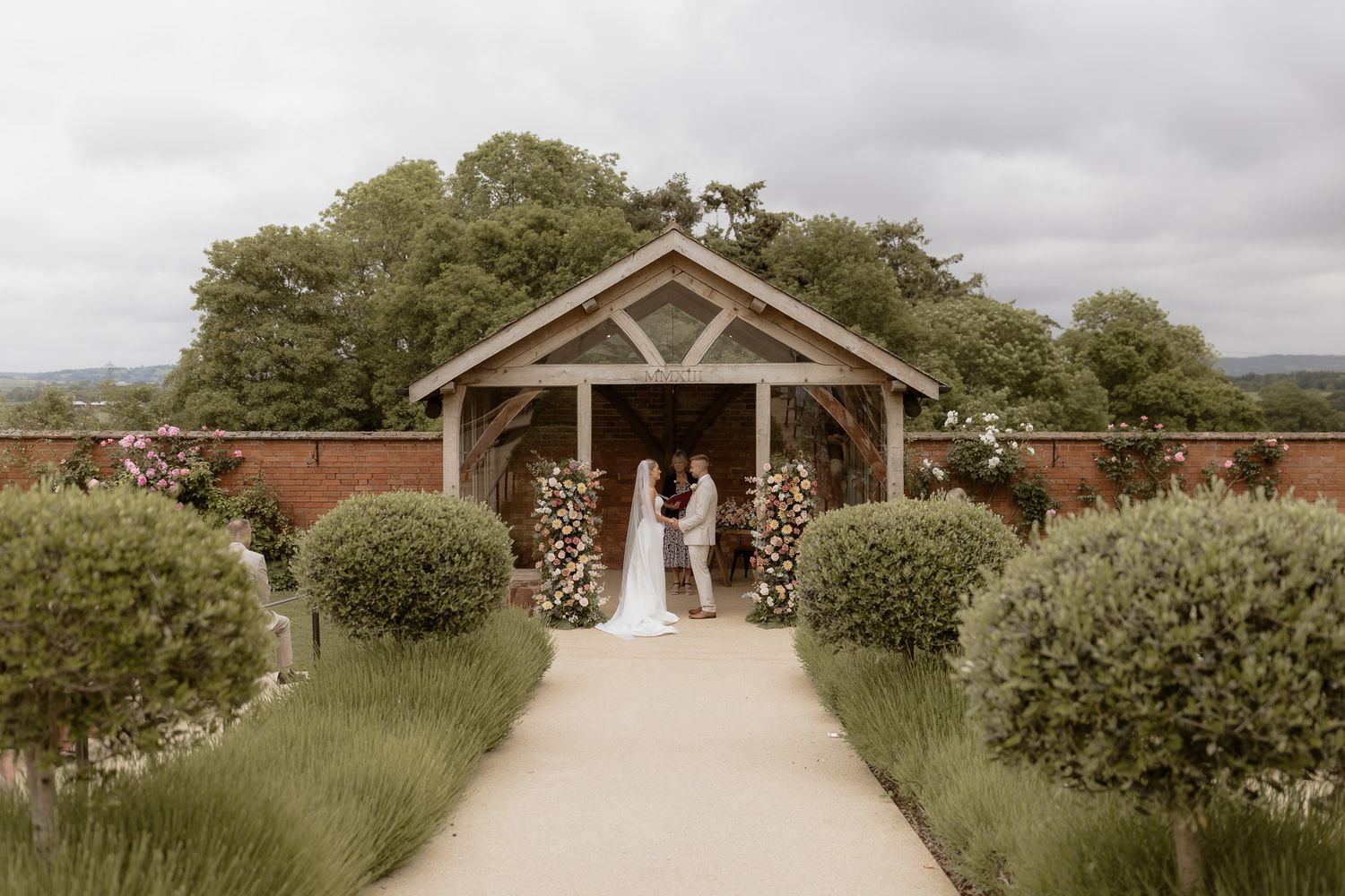 Rustic outdoor wedding ceremony under a wooden pavilion with floral arrangements and brick wall backdrop.