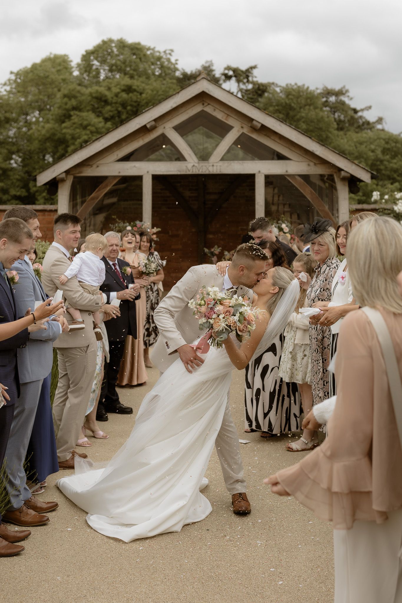 Newlyweds share romantic kiss under wooden pavilion surrounded by wedding guests.