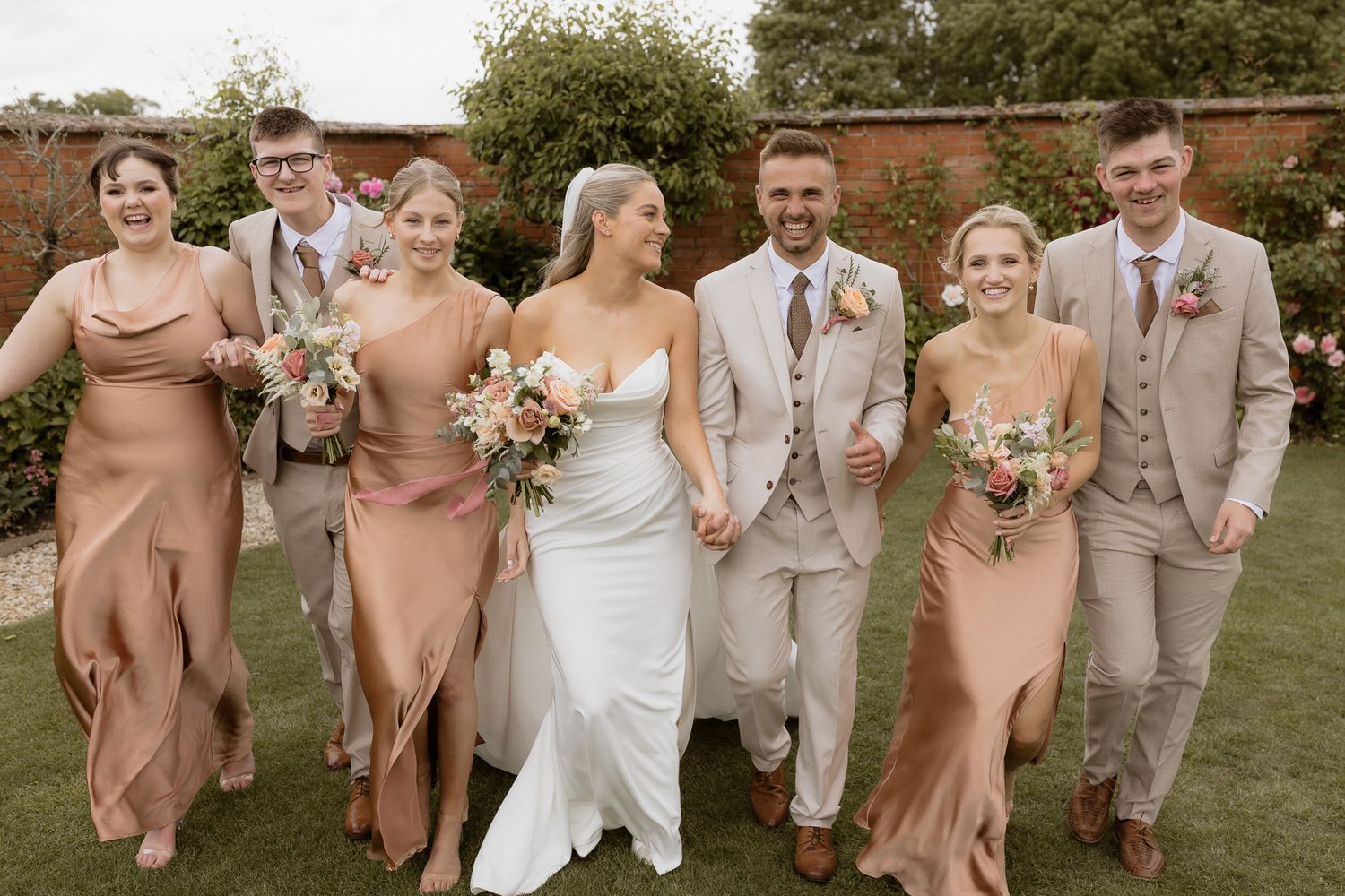 Wedding party in beige suits and blush dresses posing together on green lawn.