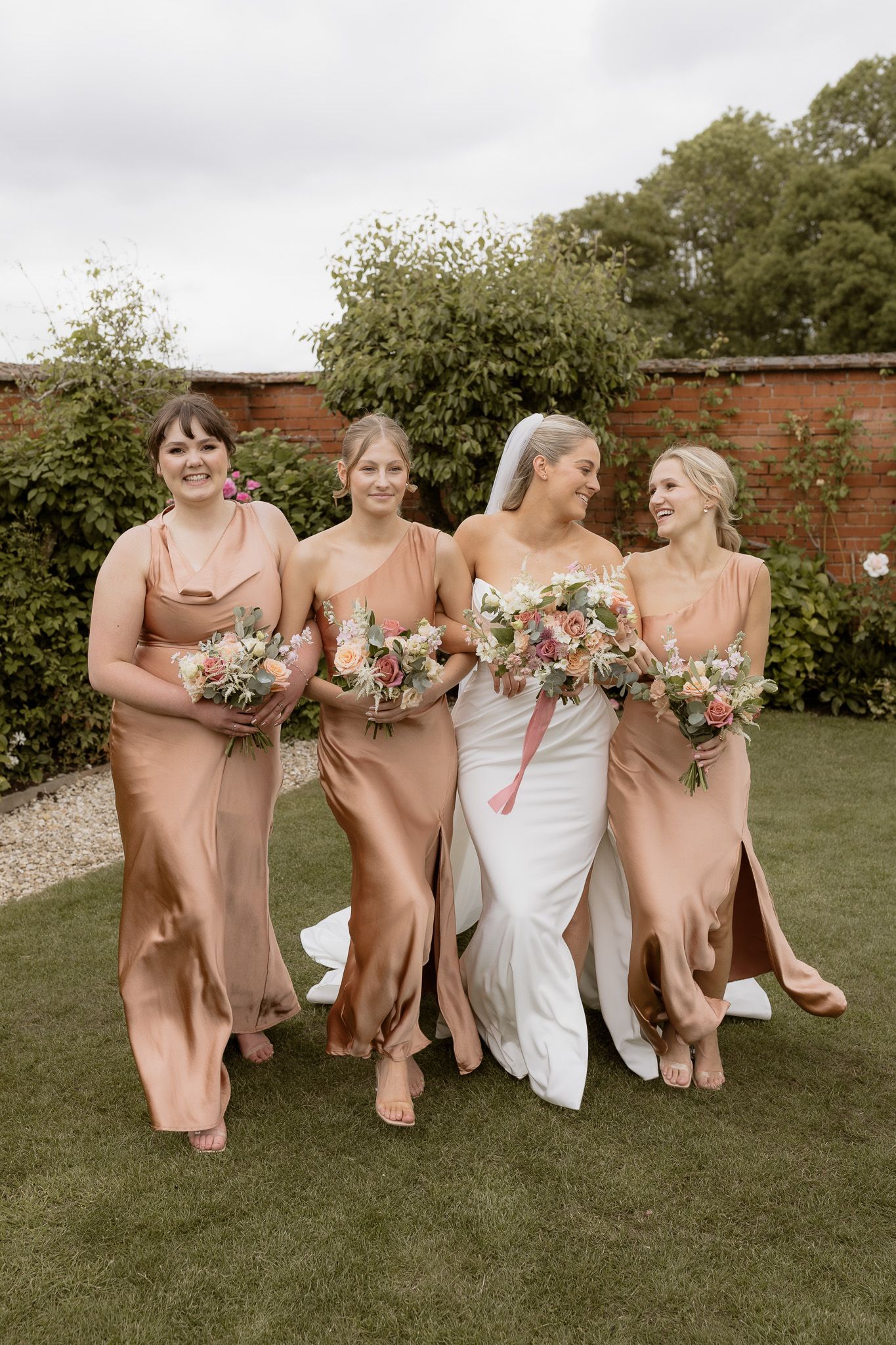 Bridal party in blush silk dresses walking together on grass holding floral bouquets.