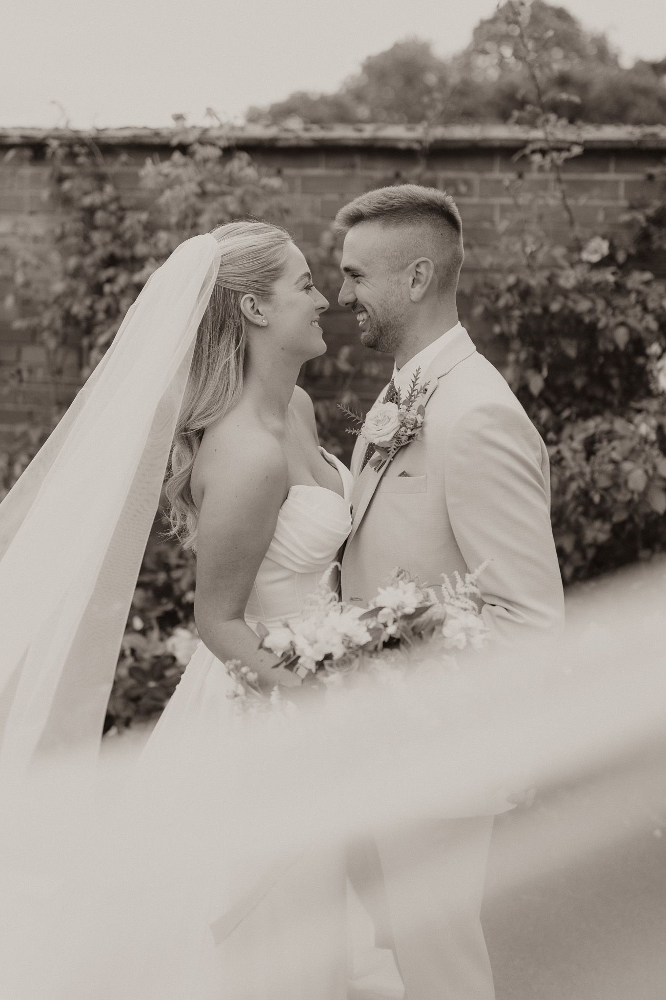 Romantic black and white portrait of newlyweds embracing by brick wall.