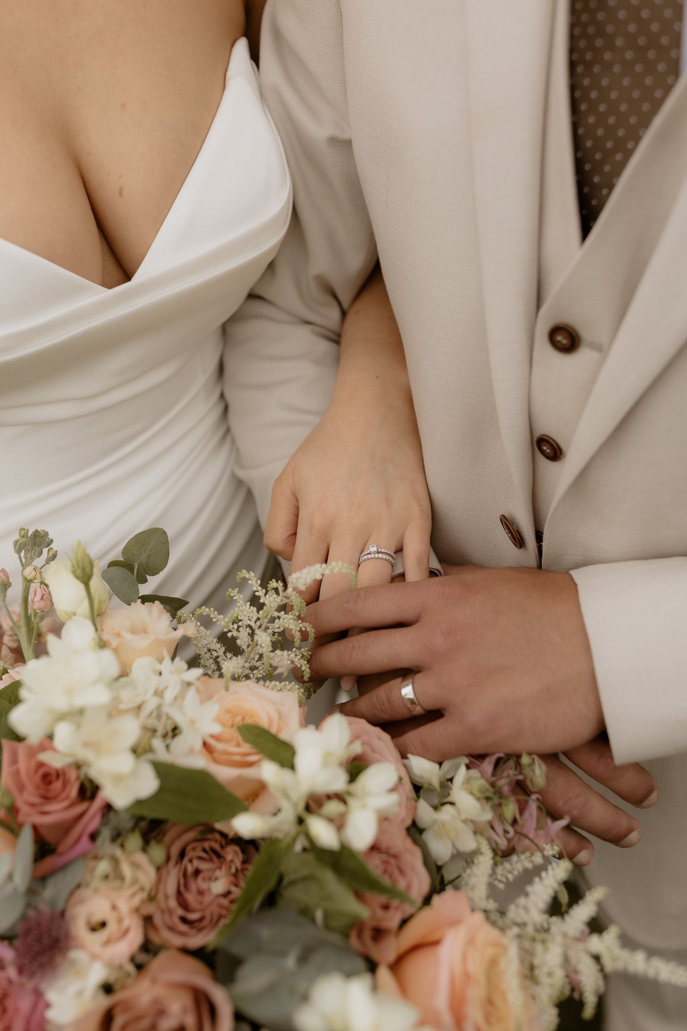 Close-up of wedding couple's hands with rings next to a romantic pastel floral bouquet with roses and greenery.