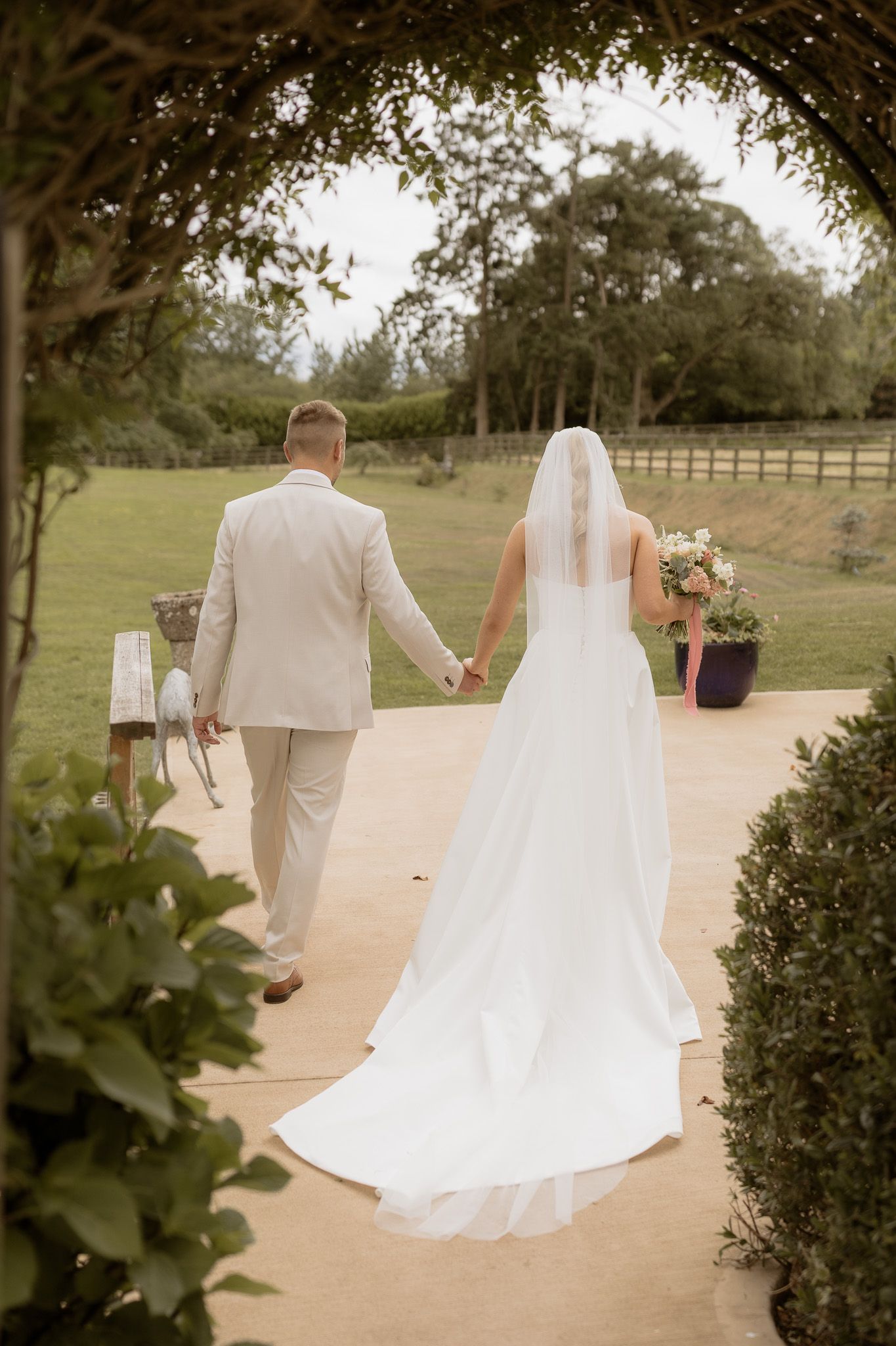 A couple walks hand in hand down a garden path, the bride in a flowing white gown with long train and veil.