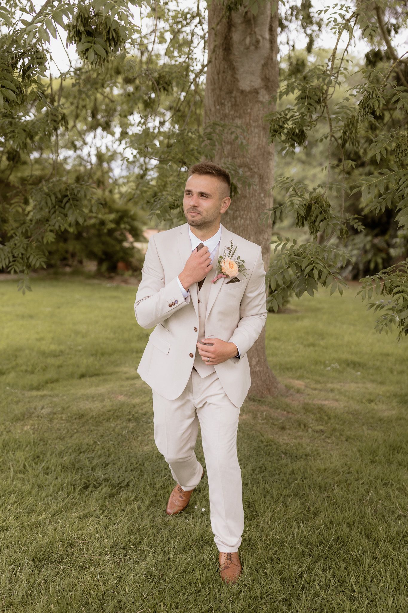 Groom in light beige suit poses confidently in garden setting.