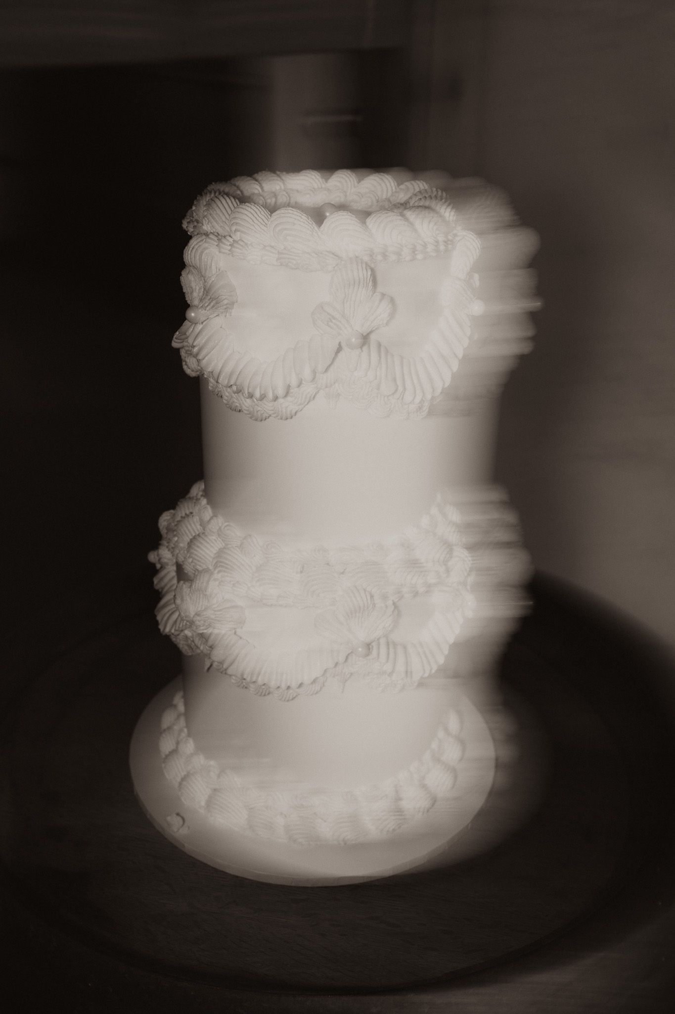 A classic white wedding cake with three tiers decorated with delicate ruffled frosting details in a soft black and white photo.