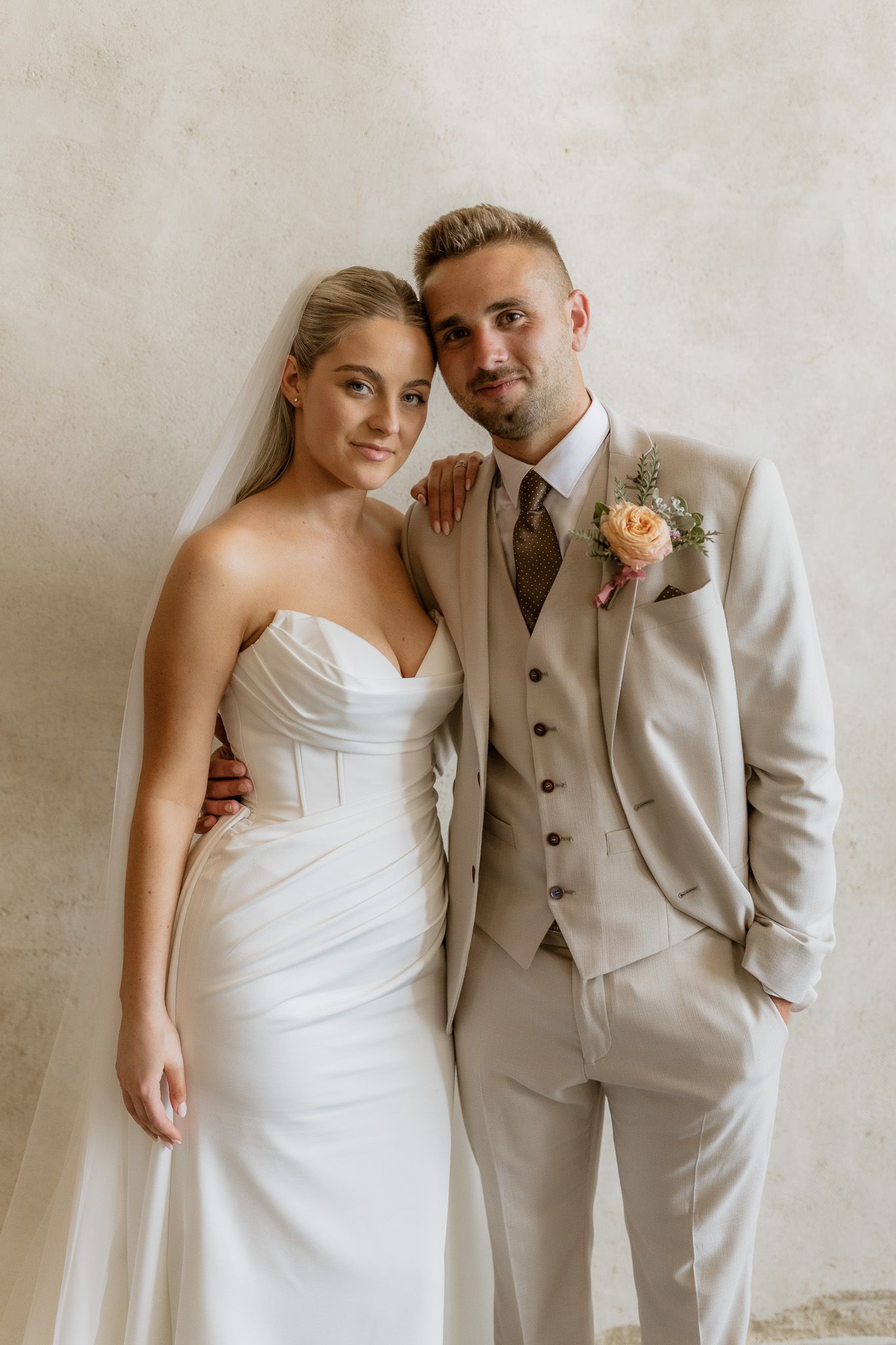 Wedding couple poses against neutral wall in elegant white dress and beige suit with peach boutonniere.