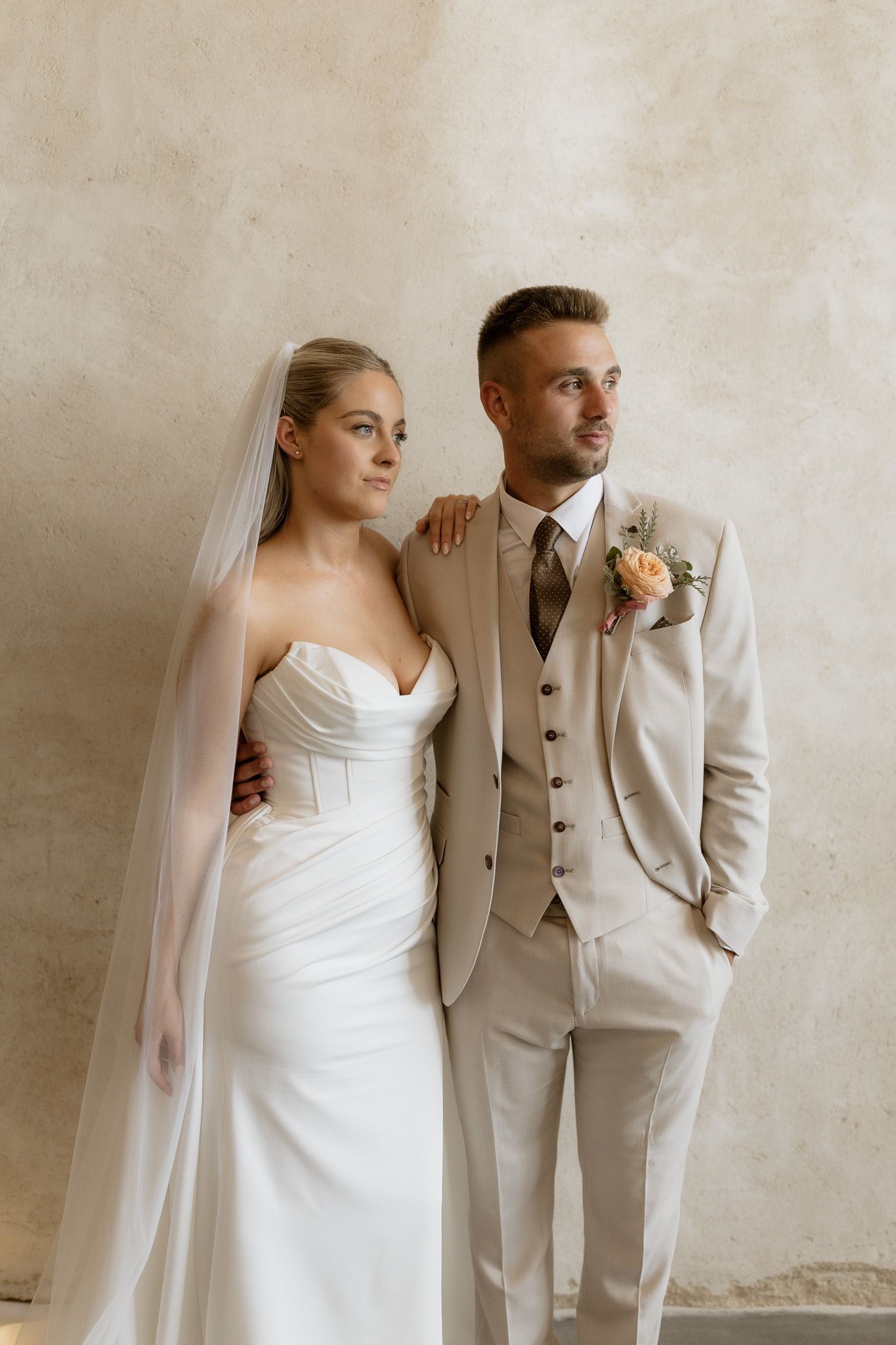 A wedding portrait of a couple in elegant attire, with the bride in white and groom in beige against a neutral backdrop.