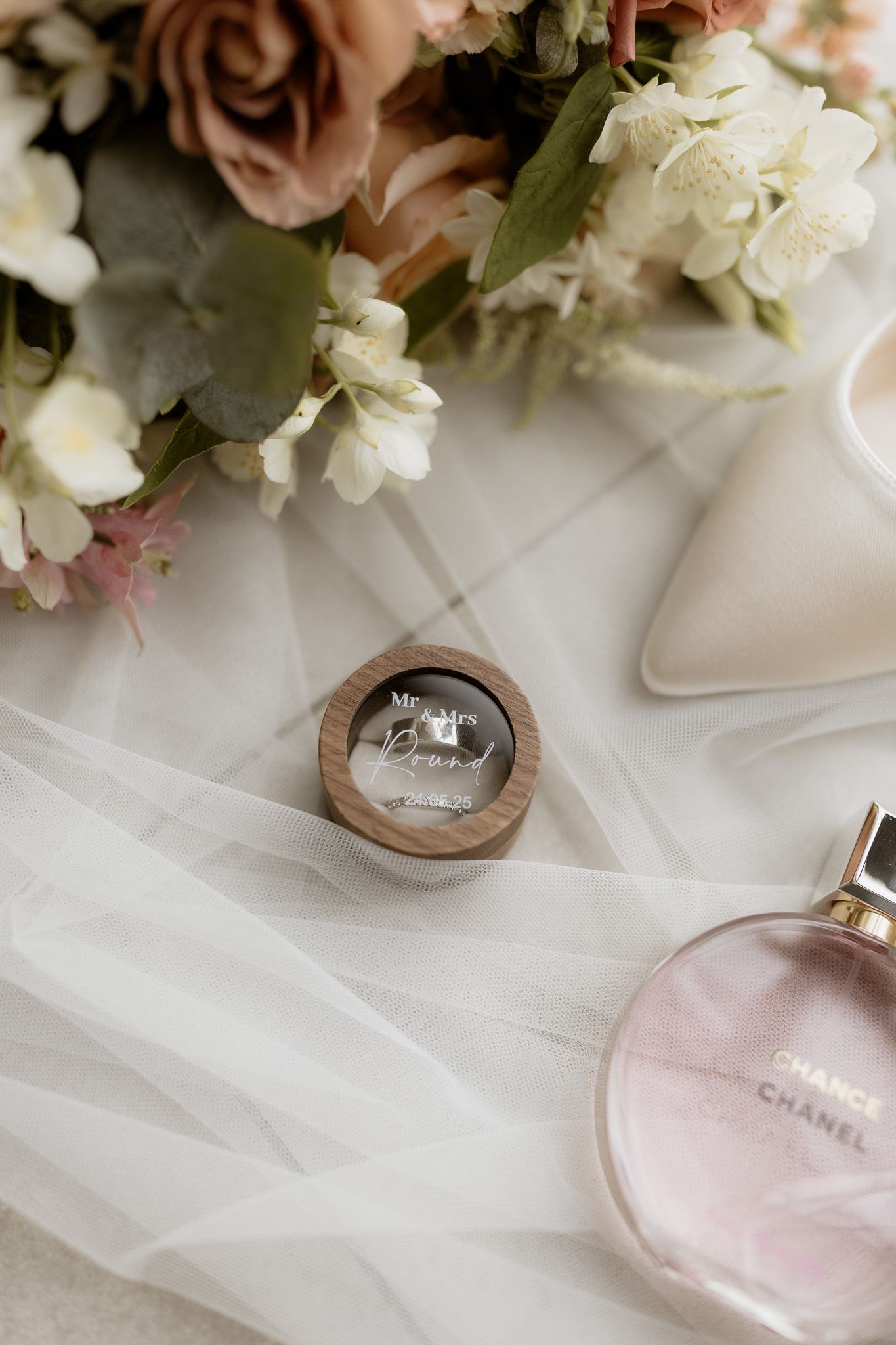 Wedding rings and Chanel perfume bottle arranged with delicate roses and tulle fabric on white surface.