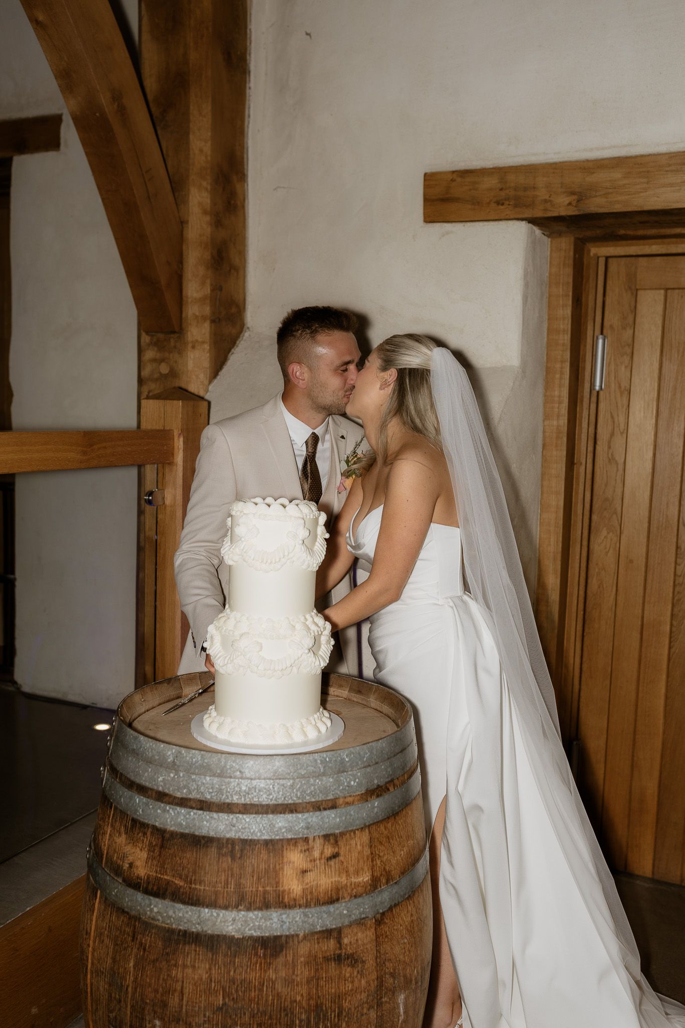 A wedding couple sharing a moment while cutting their white tiered cake displayed on a rustic wine barrel.