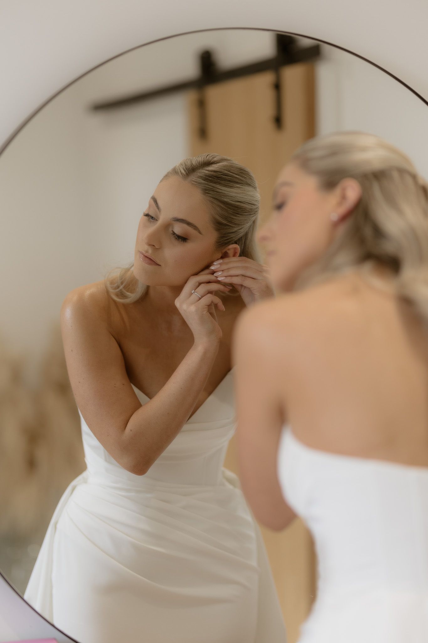 A bride in a strapless wedding gown putting on earrings while looking in a round mirror.