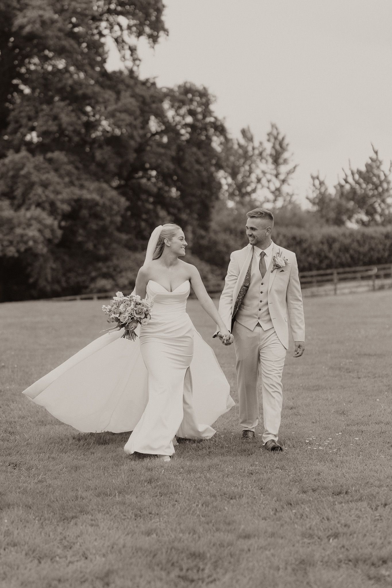 A black and white photo of newlyweds walking hand in hand across a grassy field with trees in the background.