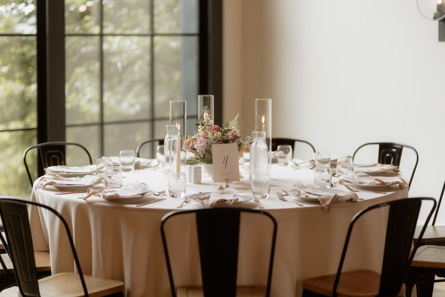 An elegant wedding reception table set with white linens, vintage metal chairs, and delicate floral centerpieces in soft natural light.