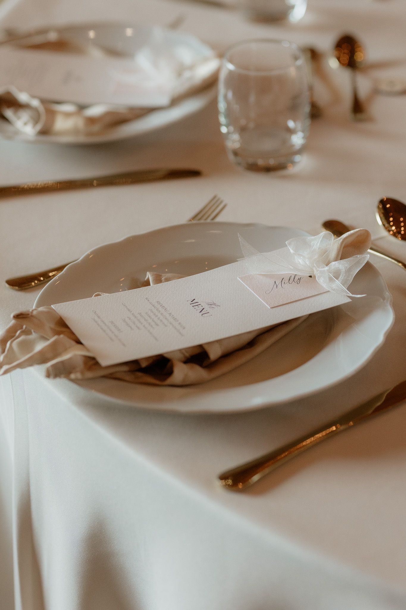 Close-up of an elegant place setting with white plates, menu card and silverware on crisp white tablecloth at wedding reception.