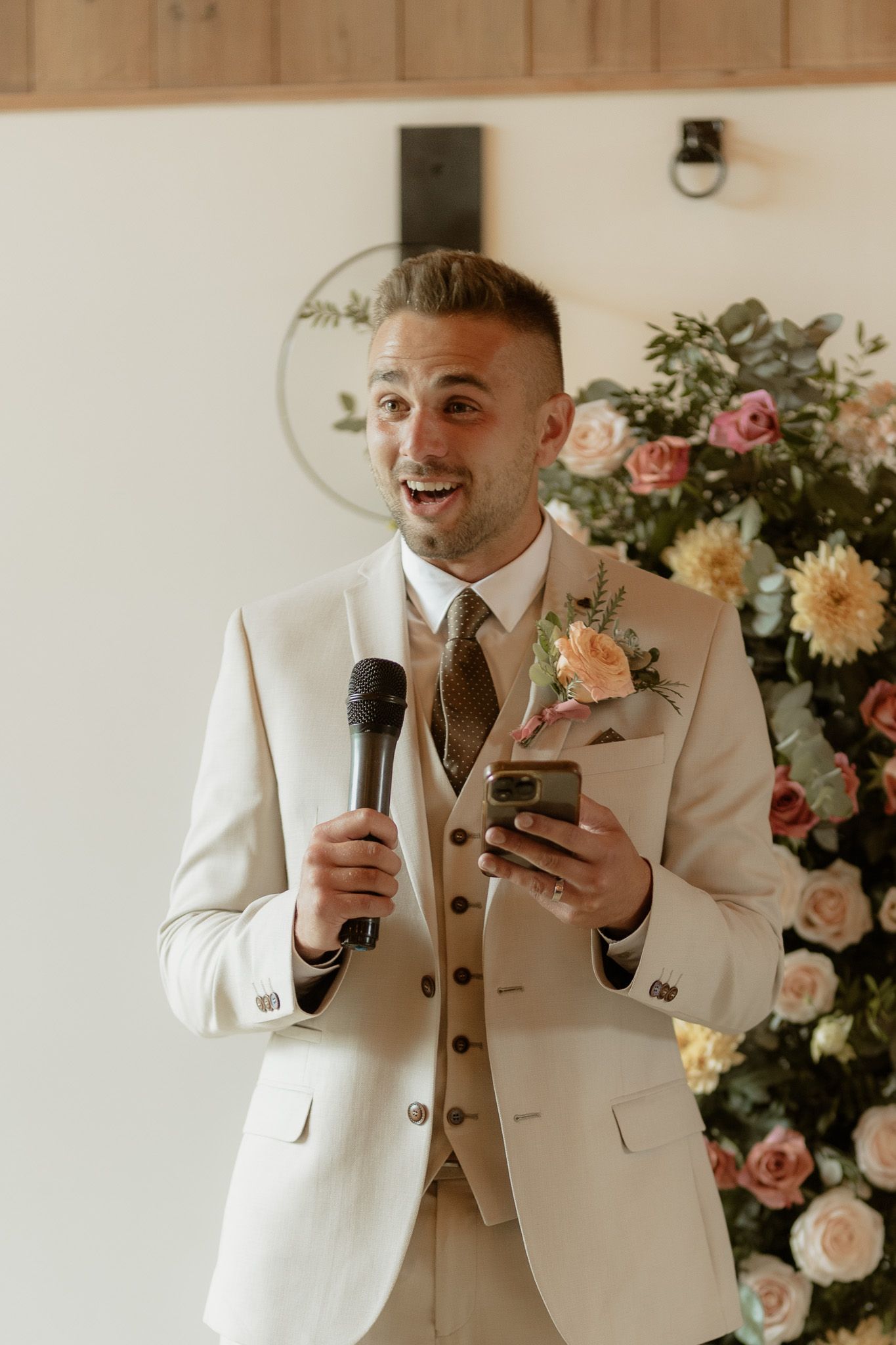 Wedding speech at reception with tan suit and floral decor against white wall and pink roses.