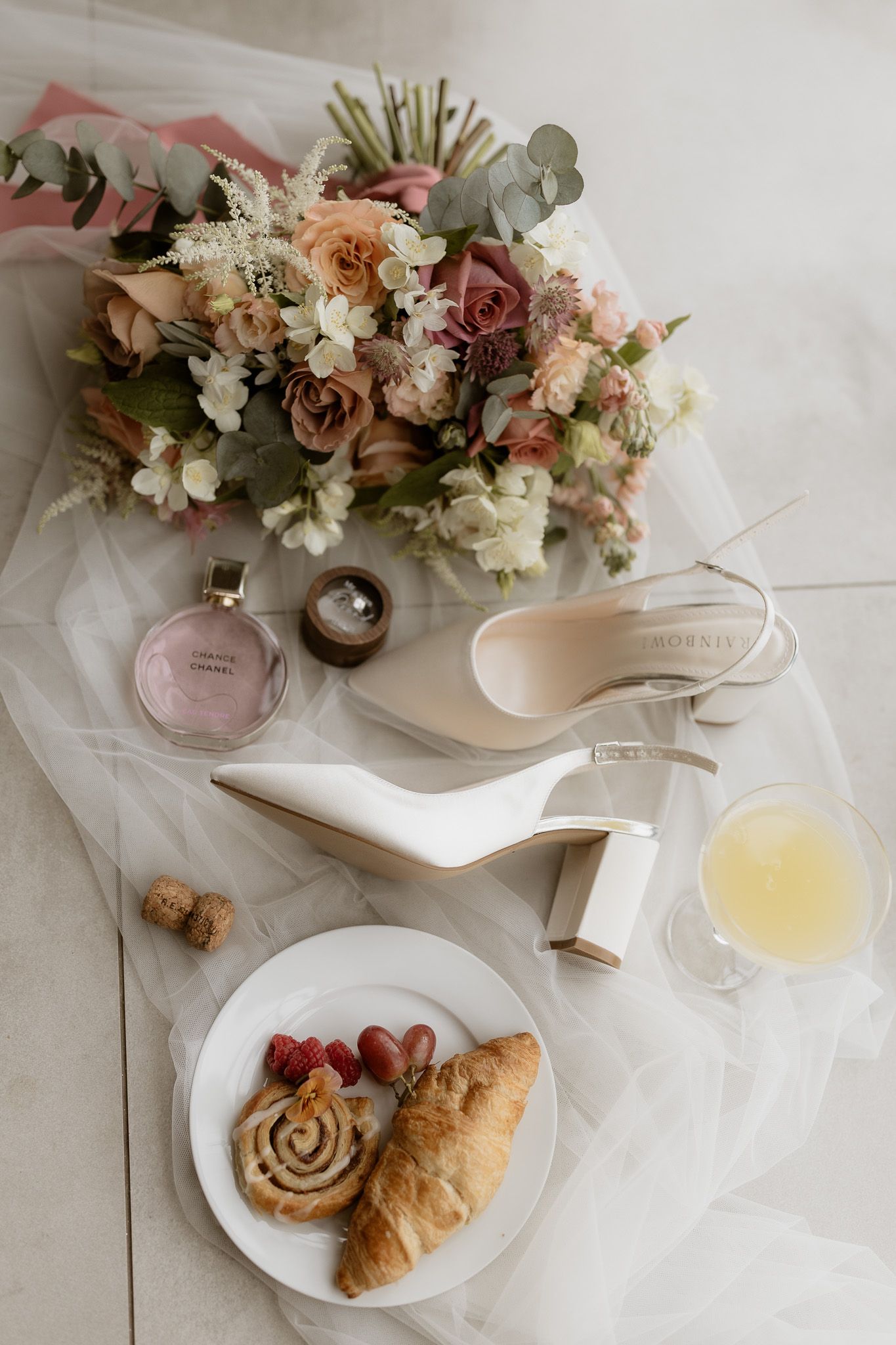 Wedding details flatlay with white heels, floral bouquet, perfume, and pastries on white fabric.