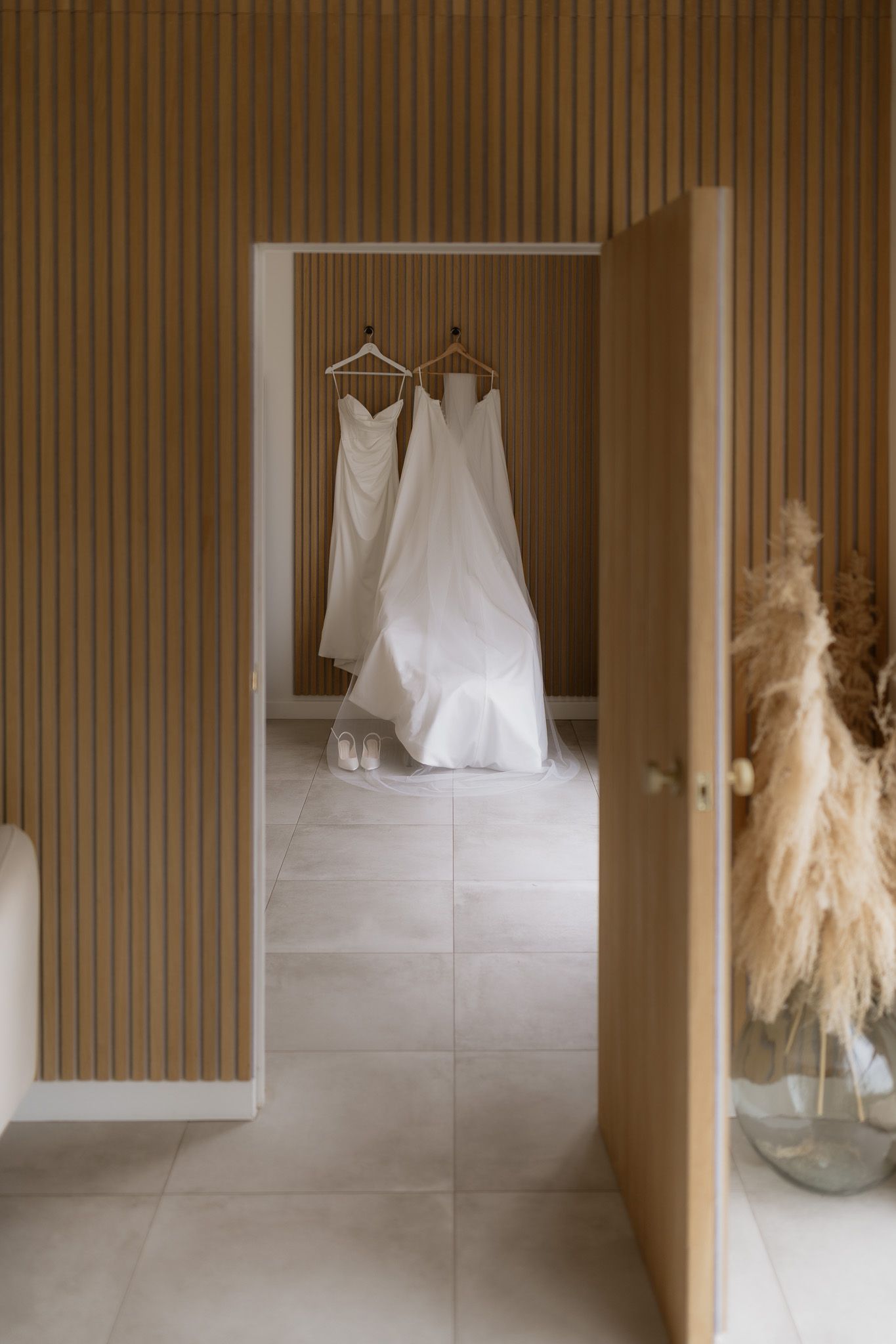 Two elegant white wedding dresses hanging in a doorway with light wood paneled walls and neutral tiled floors.