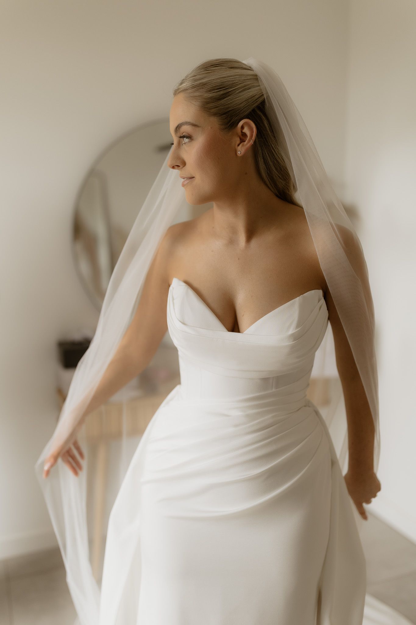 A bride in an elegant strapless white wedding dress and veil stands in a minimalist white room.