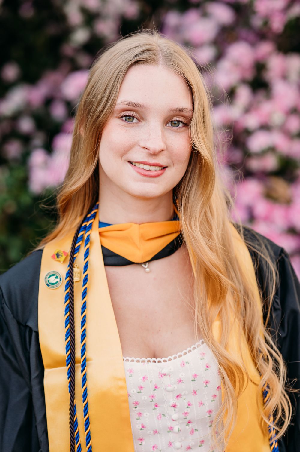 Graduate in black cap and gown with gold honors stole smiles in front of pink cherry blossoms on graduation day.