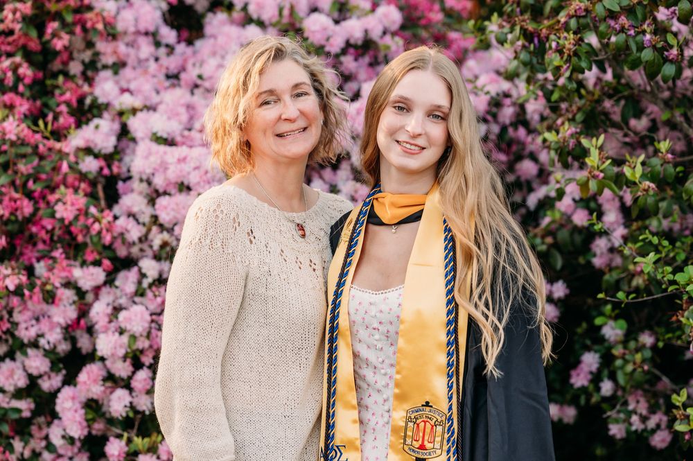 Two people smiling together in front of blooming pink azalea bushes during a graduation celebration.