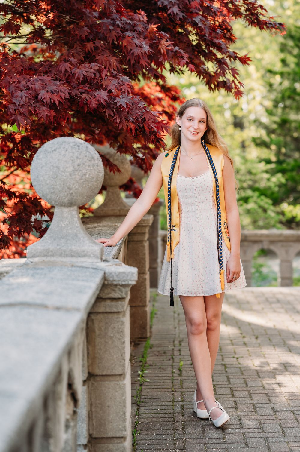 A graduate in a beige dress and gold stole stands near stone architecture with vibrant red Japanese maple foliage overhead.