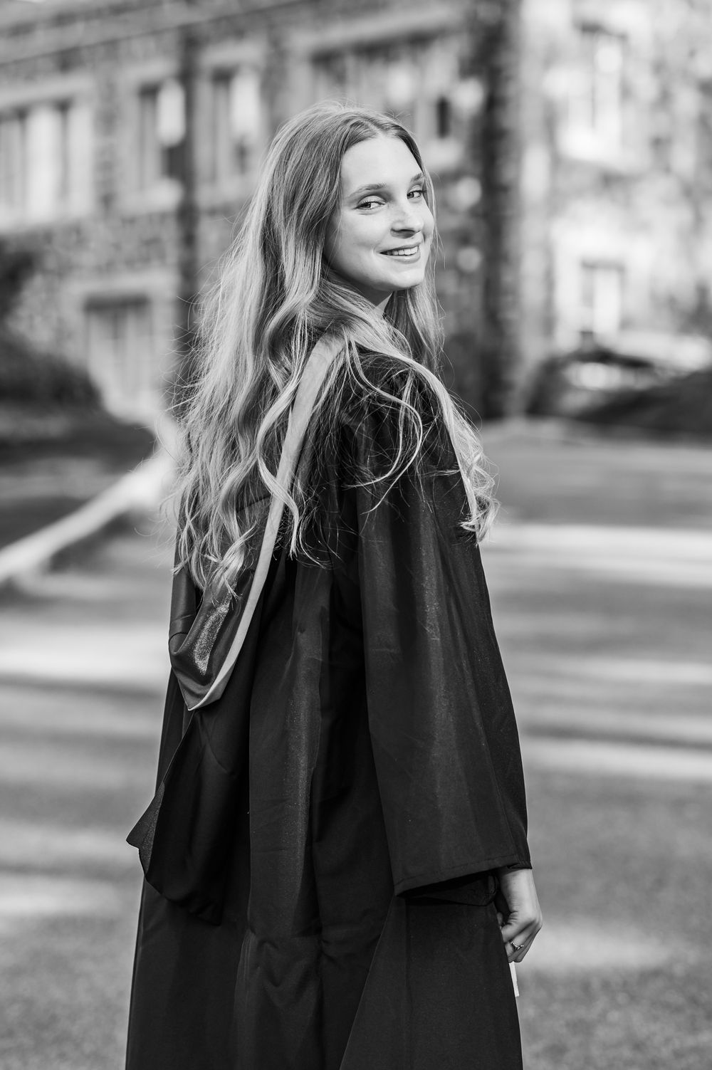 Black and white portrait of a person with long wavy hair wearing a graduation gown while looking back and smiling outdoors.