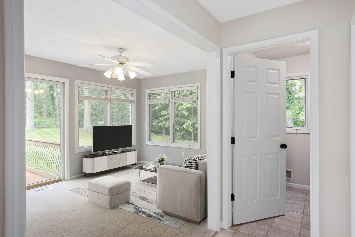 Virtually staged sitting room with neutral furniture, modern TV console, large windows, ceiling fan, and serene forest views.