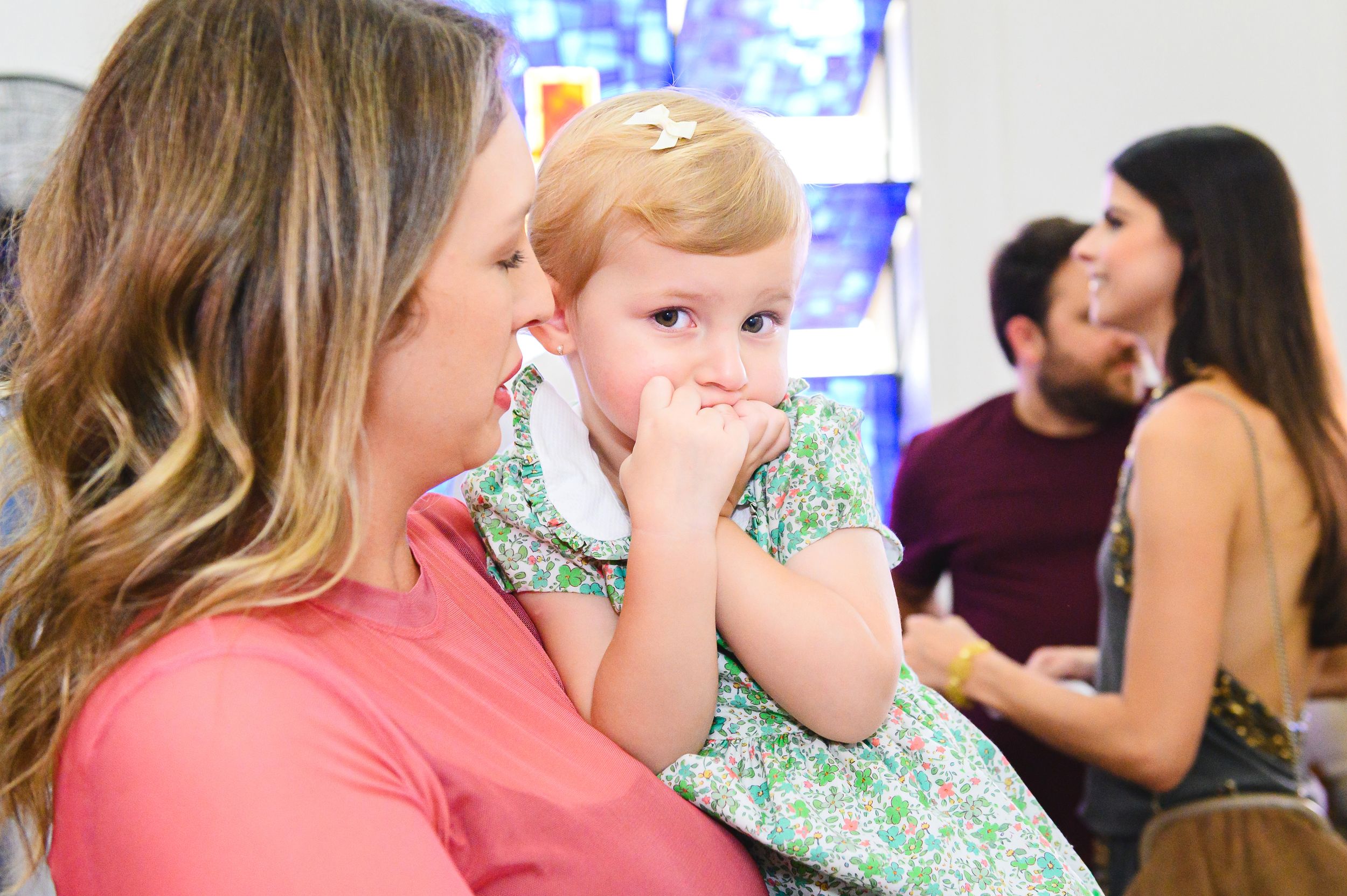 A parent and child in pink and floral outfits share a whispered moment while others chat in the background.