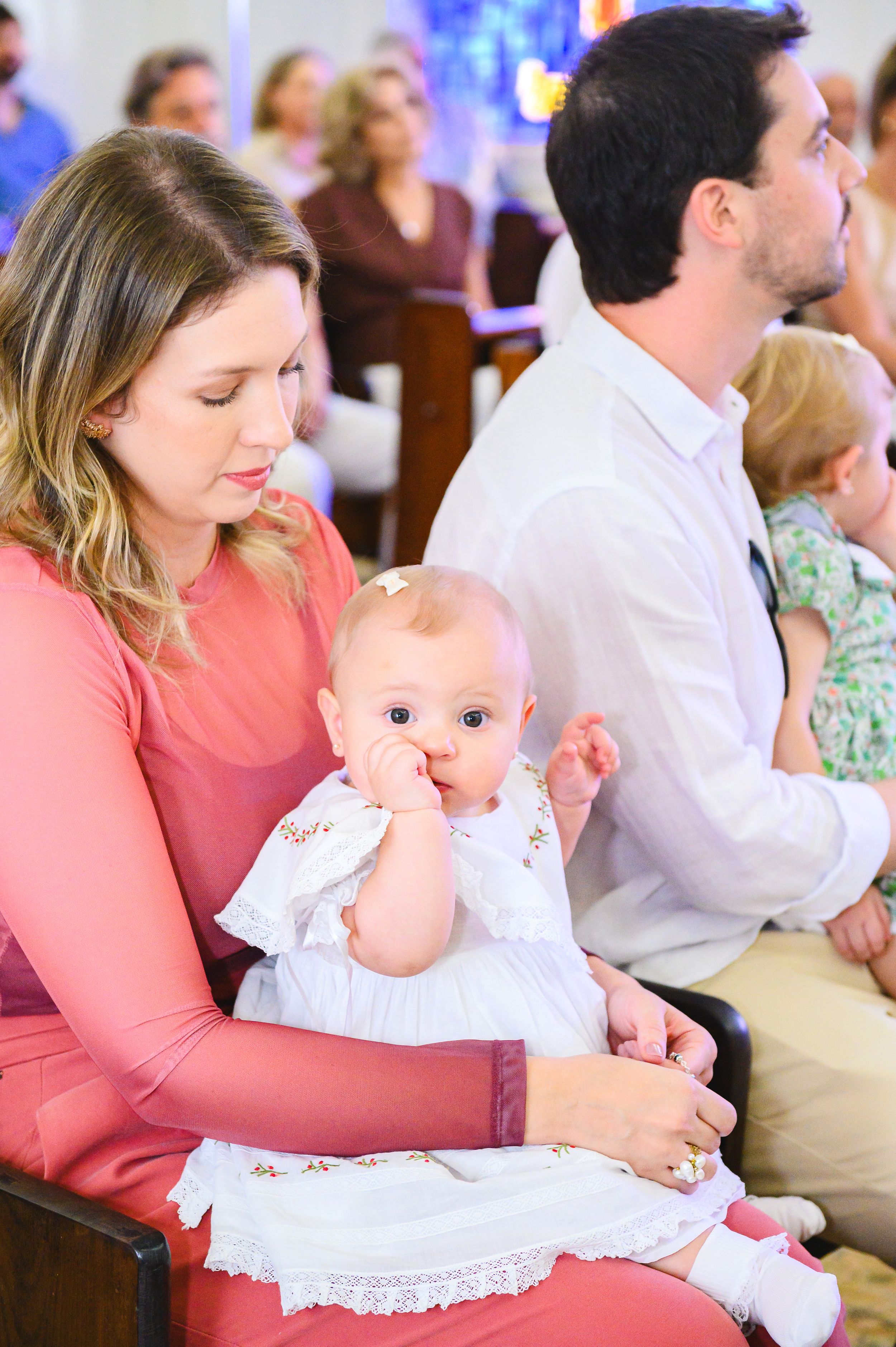 Young family attending church service with baby in traditional white christening dress sucking thumb on mother's lap.